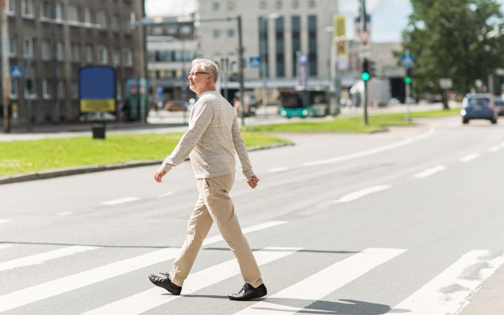 Un hombre mayor con gafas cruza por un paso de peatones en una calle urbana soleada.