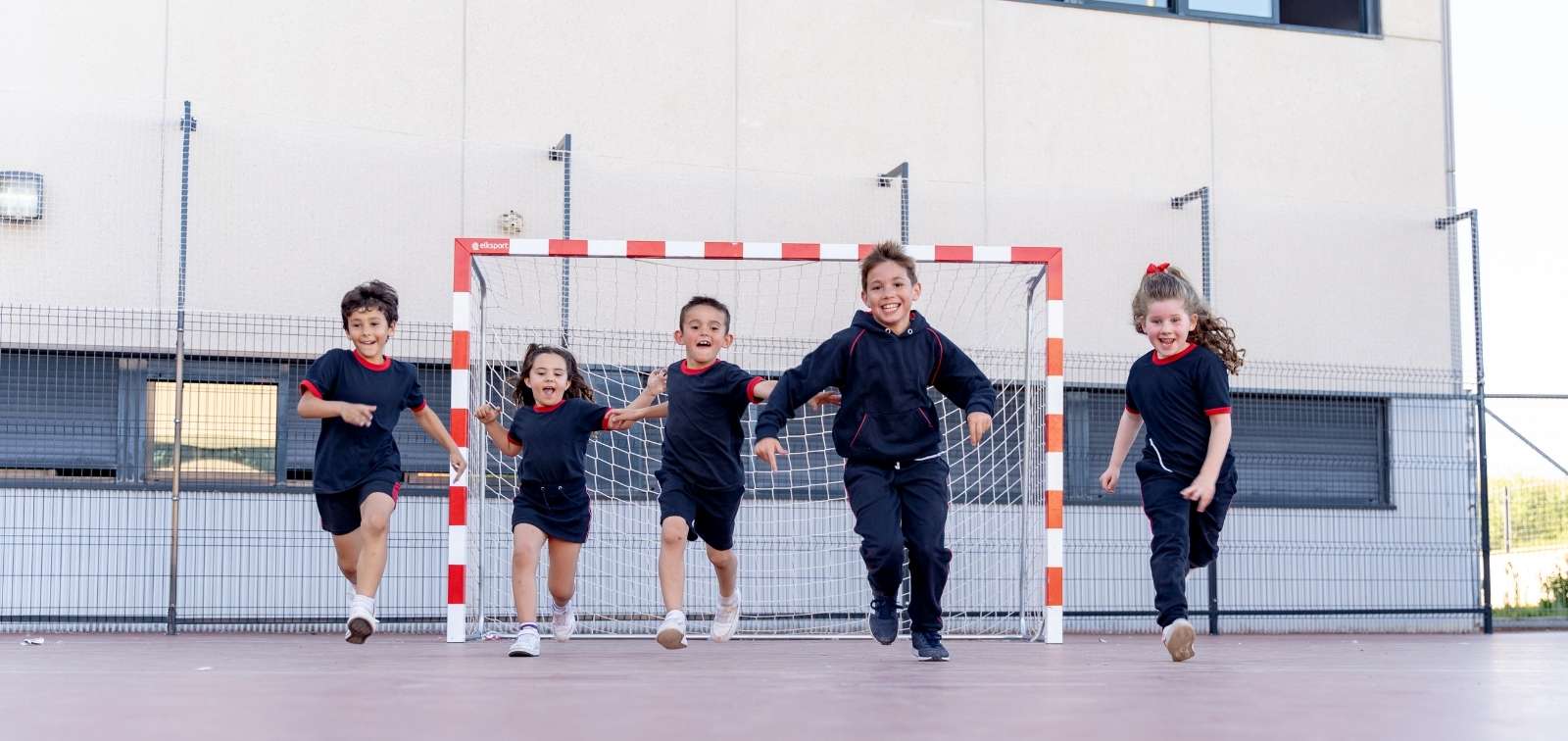 Cinco niños y niñas corriendo hacia la cámara en el patio de un colegio con una portería de fútbol de fondo.