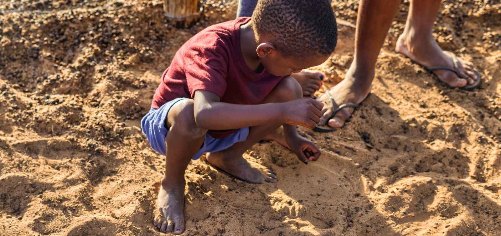 Niño africano descalzo sobre un suelo de arena y tocándola con las manos