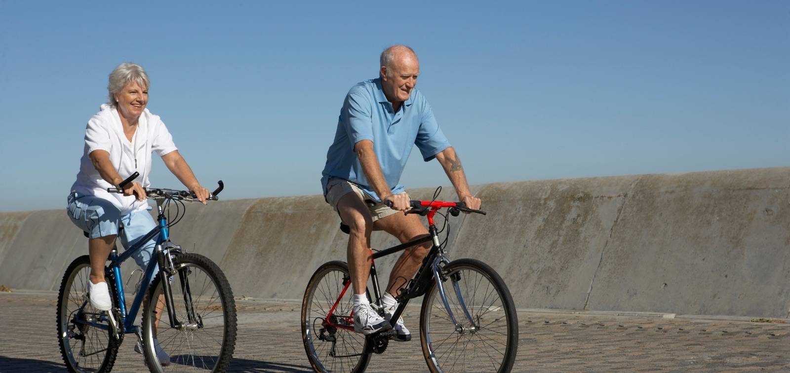 Una pareja mayor pasea alegremente en bicicleta a lo largo de un paseo marítimo en un día soleado.